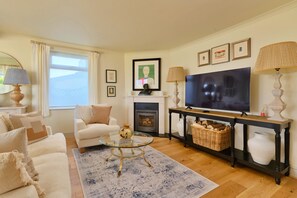 Ein Wohnzimmer mit Sofa, Sessel, Glastisch, Kamin, Fernseher und dekorativen Lampen.