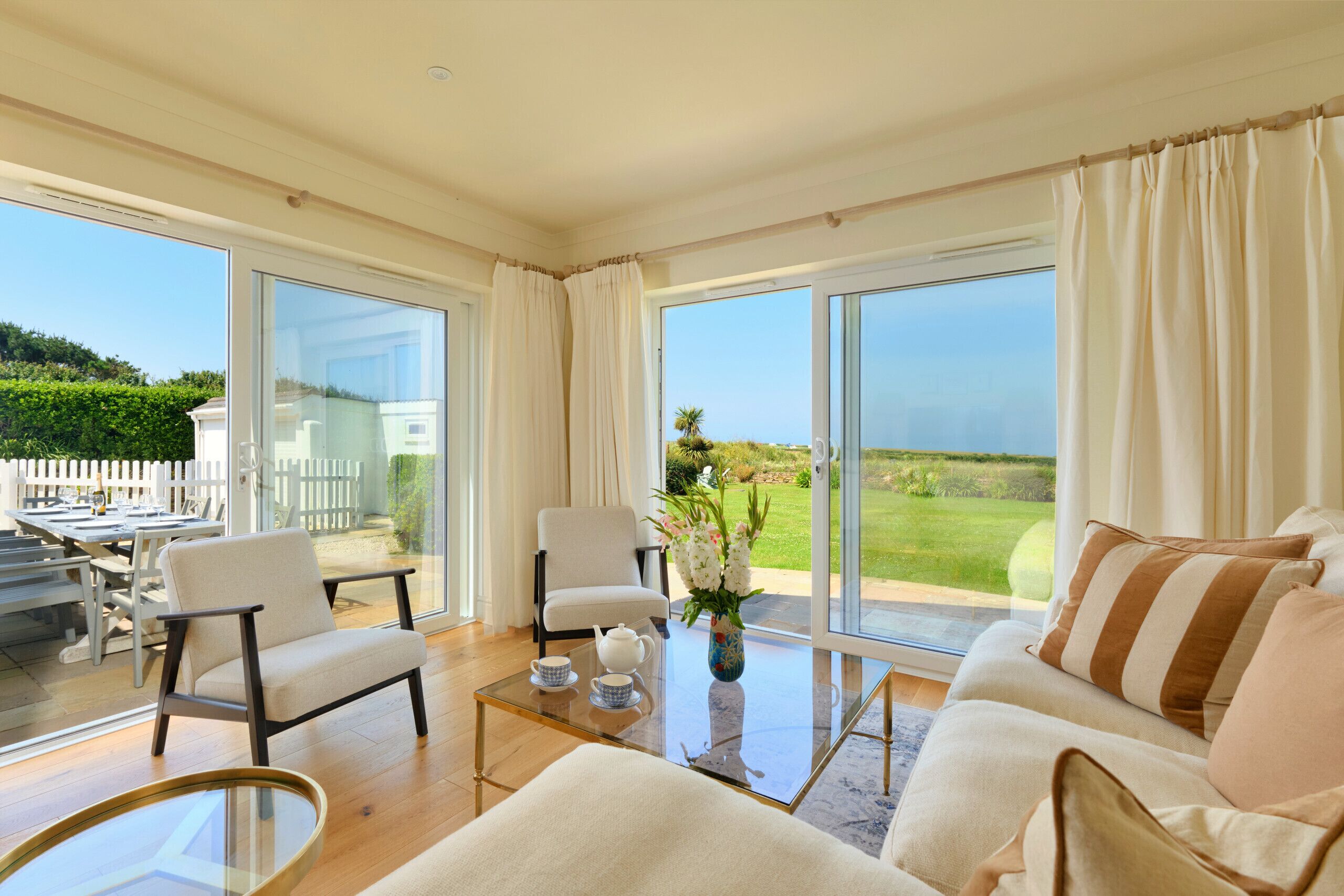 A spacious living room with a glass coffee table, a sofa, and a chair. There is a view of a green landscape through large windows.