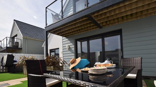 A patio with wicker furniture, a table set for a meal, and a view of a house with a balcony. - 3 Bed in Bideford (oc-40820) (Northam)