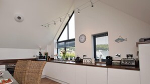 A modern kitchen with a dining area, featuring a high ceiling, large windows, and a clock on the wall. - 3 Bed in Bideford (oc-40820) (Northam)