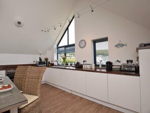 A modern kitchen with a dining area, featuring a high ceiling, large windows, and a clock on the wall. - 3 Bed in Bideford (oc-40820) (Northam)