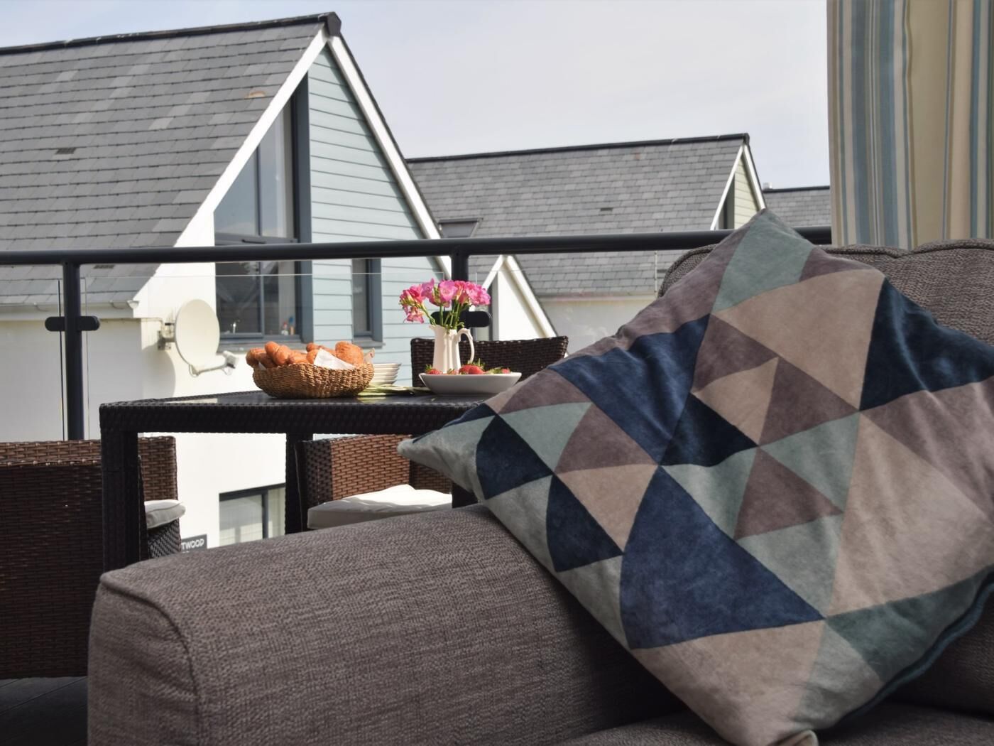 A balcony with wicker furniture, a cushion, a basket of bread, and a vase with flowers.