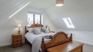 Ein Schlafzimmer mit schrägem Dach, einem Bett mit Holz-Kopfteil, zwei Nachttischen mit Lampen, einer hölzernen Bank und einem Oberlicht.