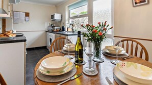 A dining table set with plates, glasses, and cutlery, a bottle of wine, and a vase with flowers in a room with a kitchen in the background. - 3 Bed in Redruth (oc-37014) (Saint Day)