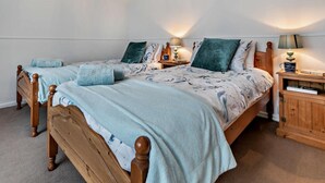 A bedroom with two wooden beds, a nightstand with a lamp, and a small wooden cabinet. - 3 Bed in Redruth (oc-37014) (Saint Day)