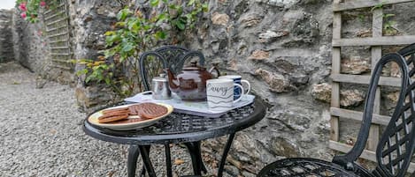 A table with a teapot, cups, and cookies, set against a stone wall with a lattice and flowering plants.