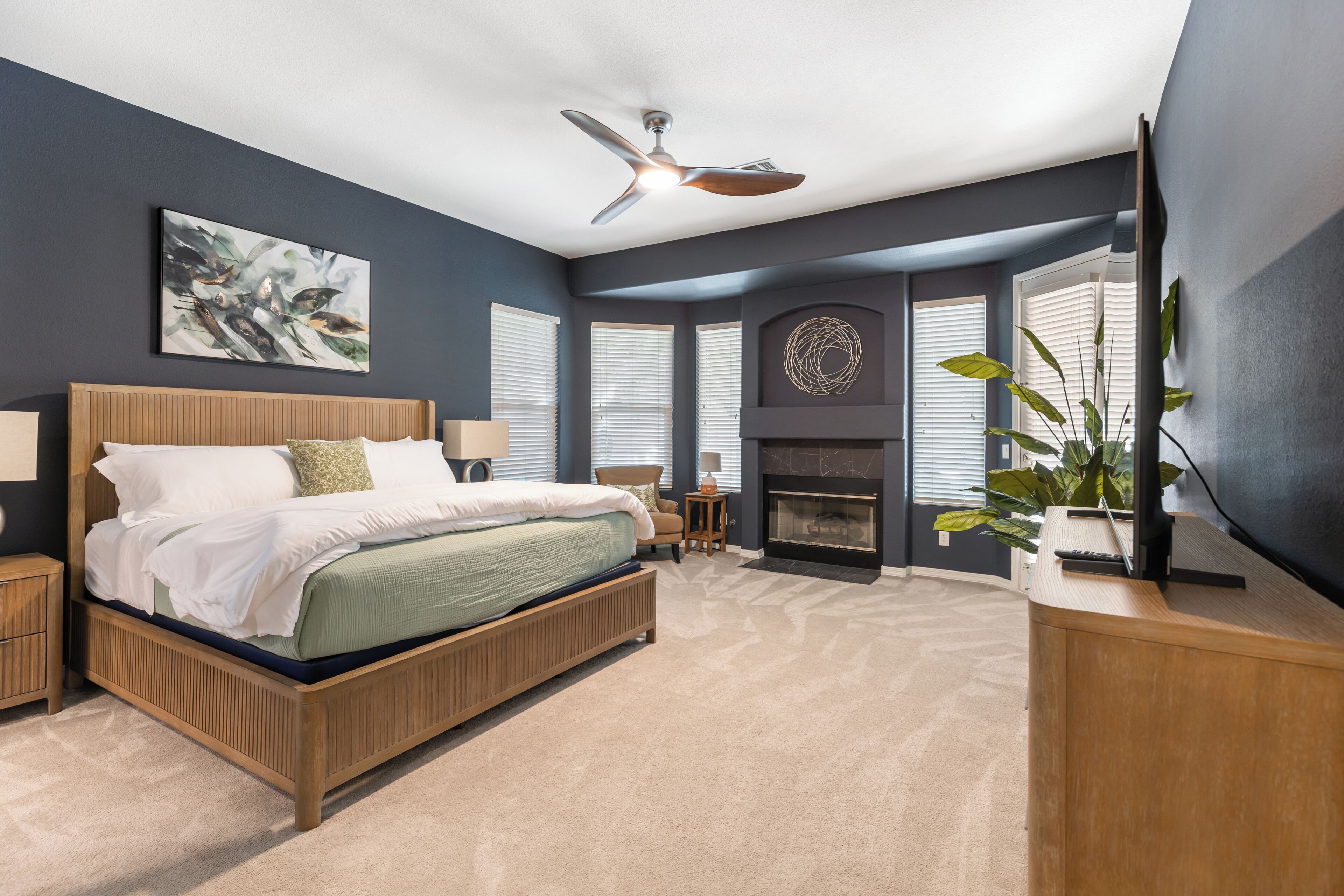 A bedroom with a bed, a ceiling fan, a fireplace, and a large window.