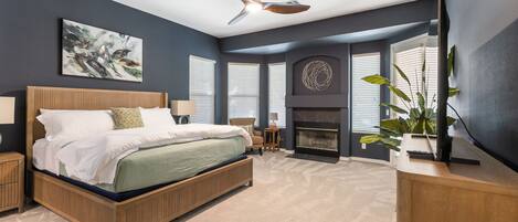 A bedroom with a bed, a ceiling fan, a fireplace, and a large window.