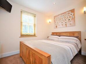 Ein Schlafzimmer mit einem hölzernen Bett, einer Bank, einem Fenster mit Jalousien, einem am Wand montierten Fernseher und einem Blumenbild.