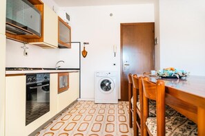 A compact kitchen with a washing machine, wooden cabinets, and a dining area with chairs. - Beautiful and Well-equipped Flat for Four (San Michele al Tagliamento)