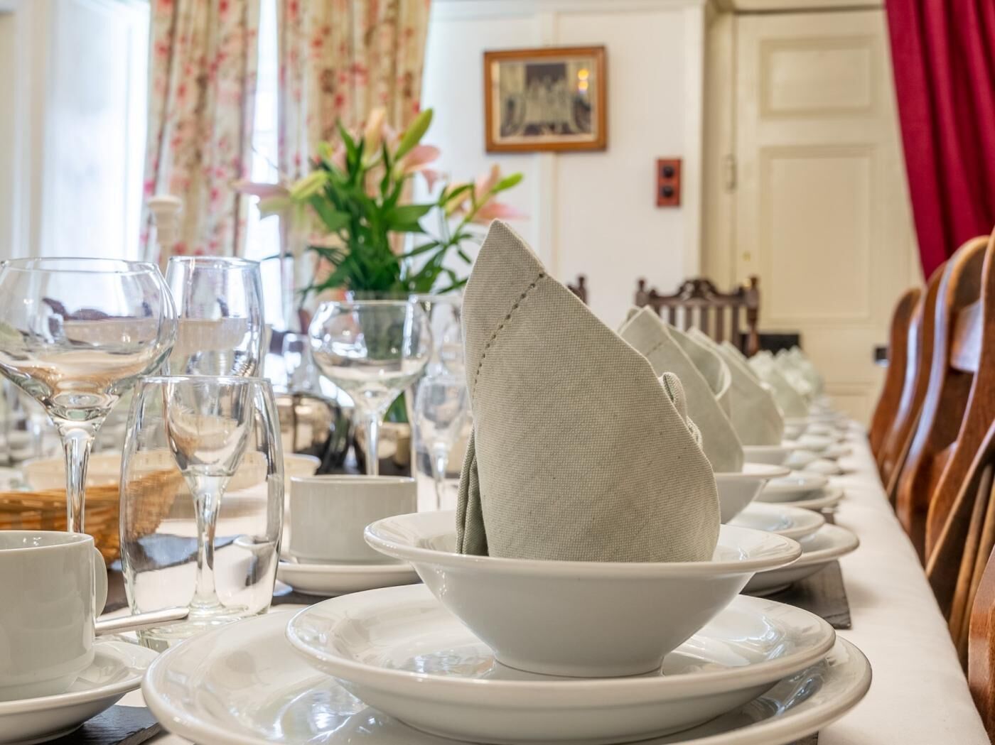 A dining table set with white plates, bowls, and wine glasses, with a vase of flowers in the background.