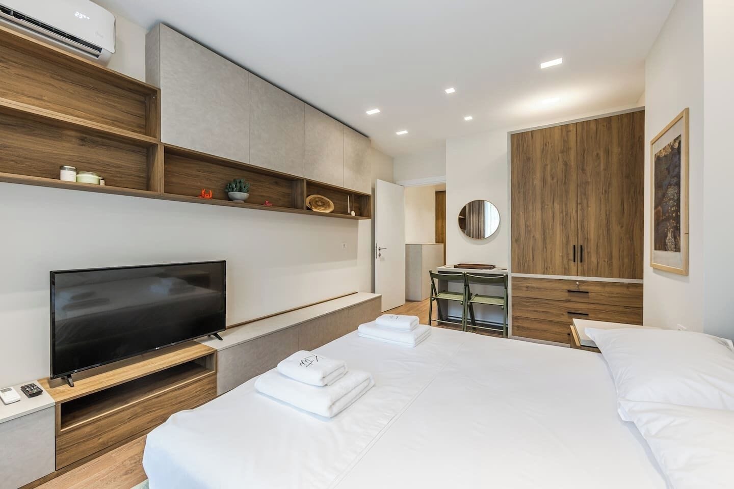 Moderni hotellihuone, jossa on taulutelevisio, valkoisilla lakanoiden peitetty sänky, puinen vaatekaappi ja pieni työpöytä tuolilla.