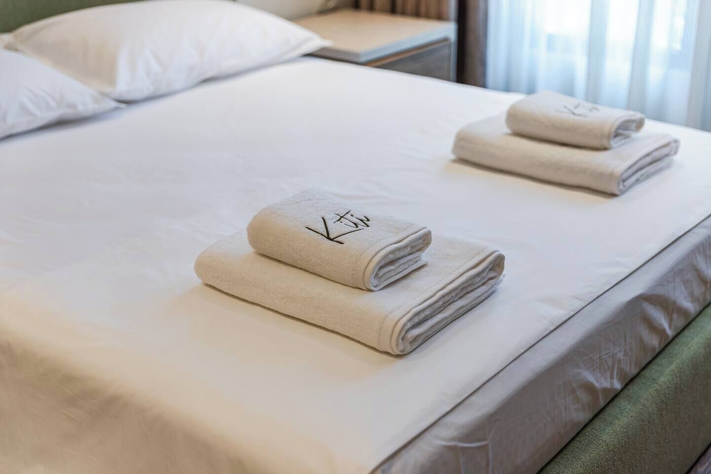 Un lit bien fait, avec du linge de lit blanc et deux serviettes pliées, chacune ornée d’un logo.