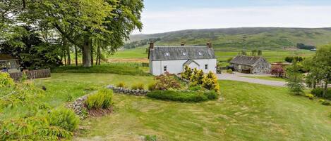 A rural house with a white exterior, surrounded by a well-maintained lawn, trees, and rolling hills in the background.