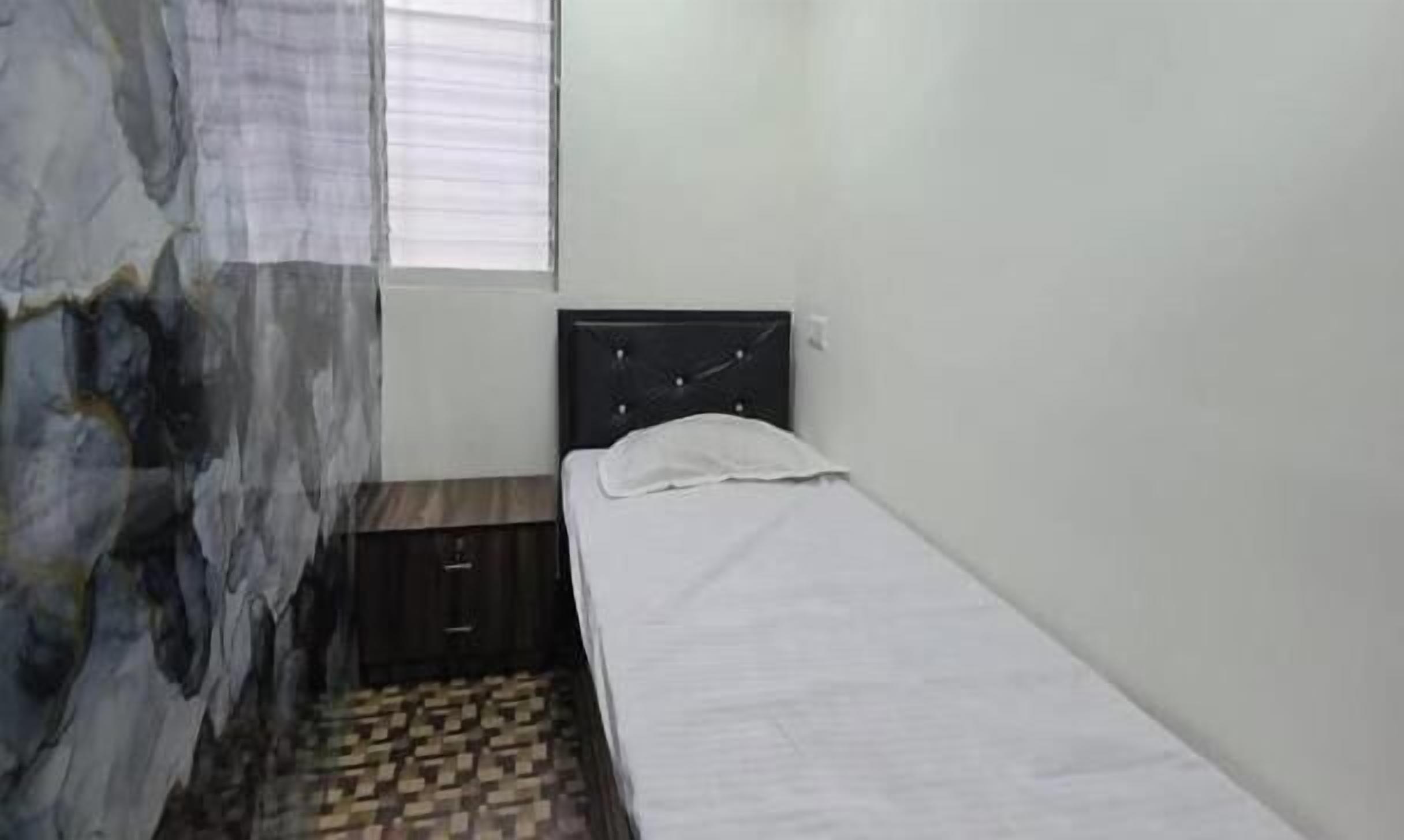 Une petite chambre avec un lit simple, un mur blanc uni et une fenêtre avec des stores.
