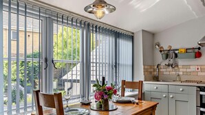 A dining area with a table set for two, a vase of flowers, and a view through large windows to the outside. - 1 Bed in Bridport (oc-37351) (Bothenhampton)