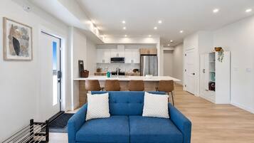 A modern living room with a blue sofa, a dining area with a white table, and a kitchen with white cabinets.