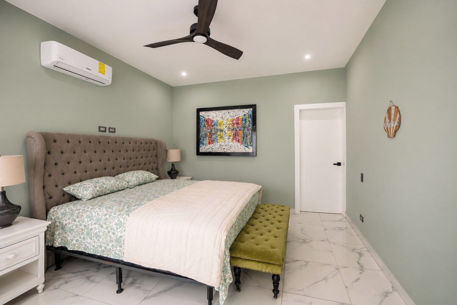 A bedroom with a tufted headboard, a bed with patterned bedding, a bedside table with a lamp, a bench, an air conditioning unit, a ceiling fan, a door, and a framed abstract painting.