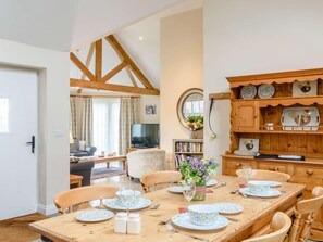 A dining area with a wooden table set for four, surrounded by wooden chairs. There is a television in the adjacent living room, and a bookshelf filled with books and a vase with flowers. - 1 Bed in Thirsk (oc-40354) (Sinderby)