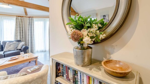 Uma sala de estar com sofá, mesa de centro, estante de livros e um espelho redondo com um arranjo floral.