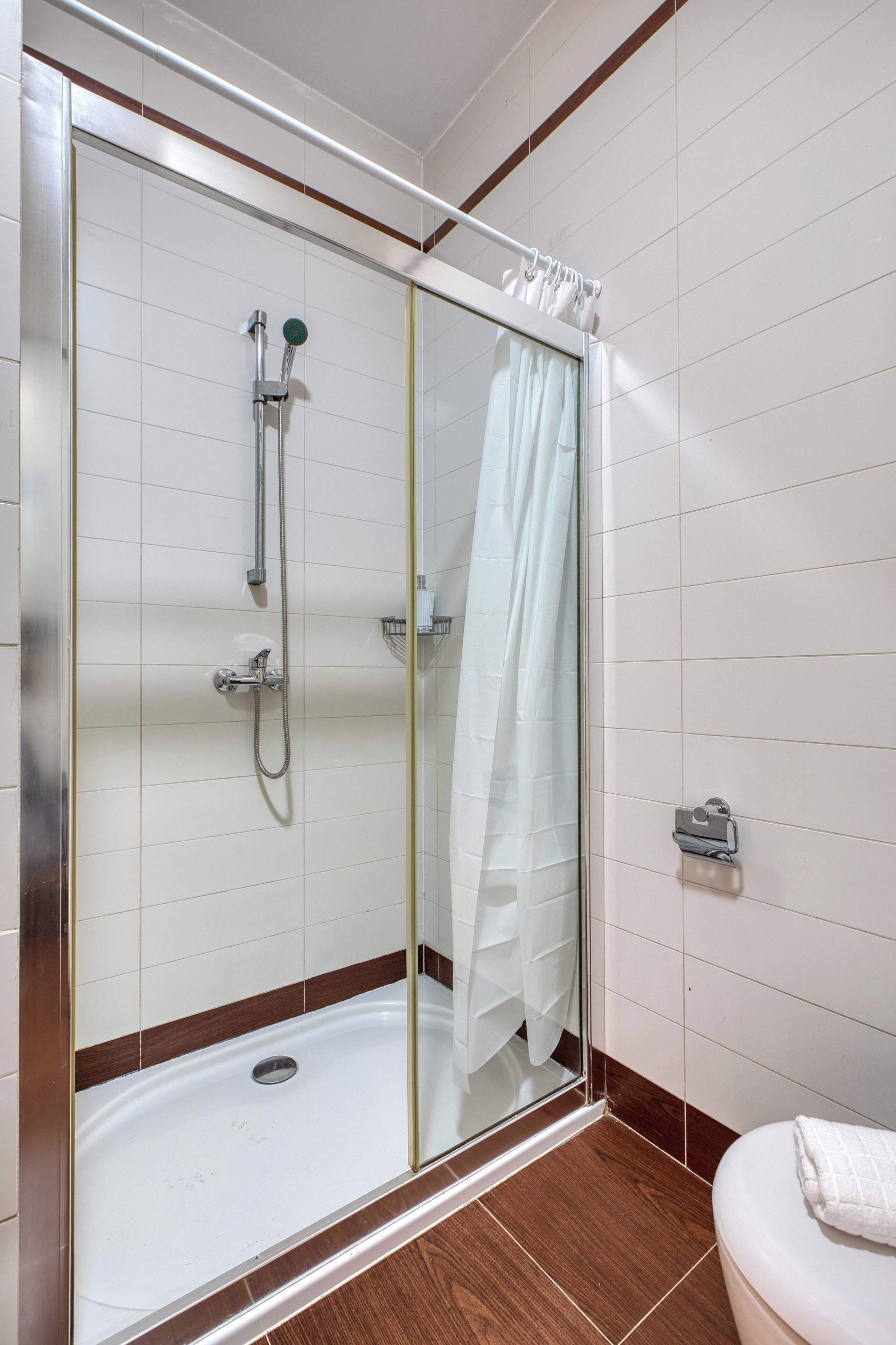 A glass-enclosed shower with a handheld showerhead and a handheld faucet, a white toilet with a towel on it, and a tiled floor with a brown border.