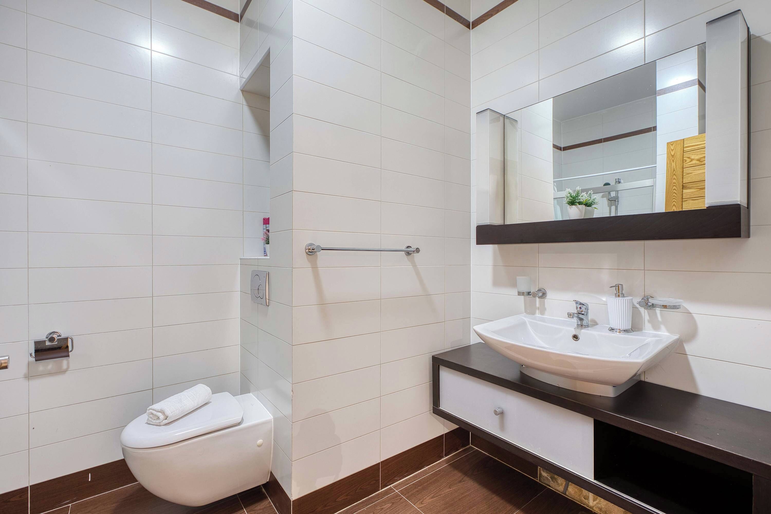 Une salle de bain moderne avec des toilettes blanches, un lavabo surmonté d’un miroir et un espace douche.