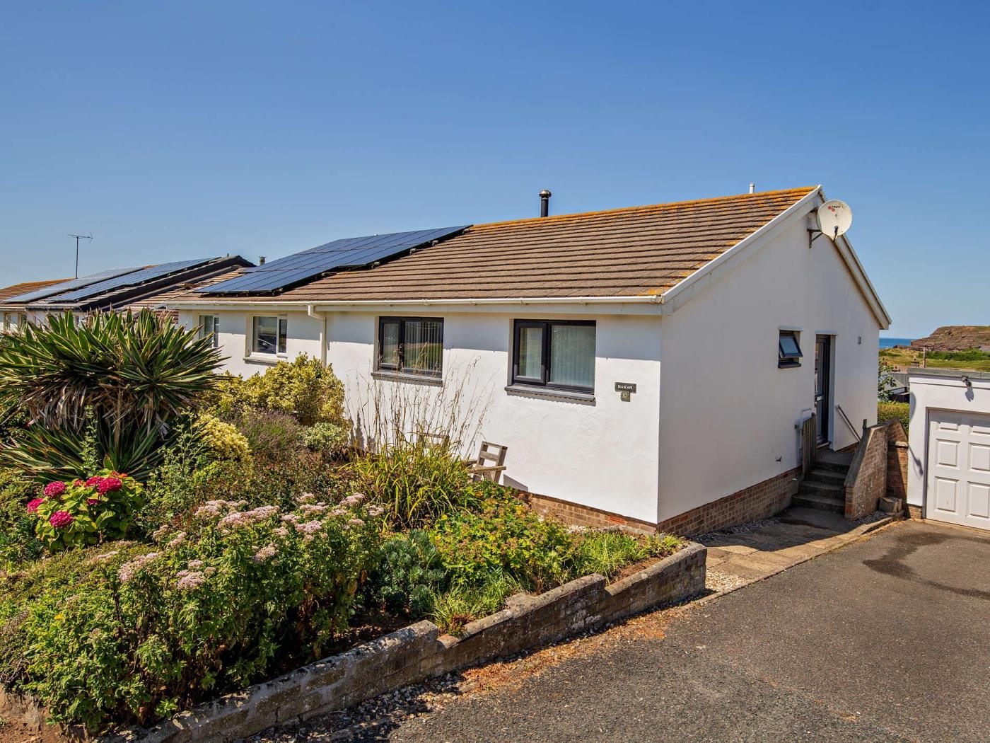 A single-story house with a sloped roof, solar panels, and a small garden with various plants and flowers.