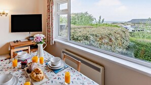 Habitación con una mesa de comedor lista para el desayuno, un televisor en la pared y un ventanal con vistas a zonas verdes y edificios.