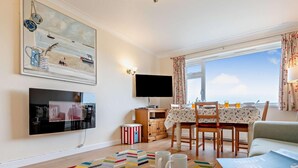 Una sala de estar con televisor, una mesa de comedor con sillas, un tapete colorido y una ventana con cortinas.