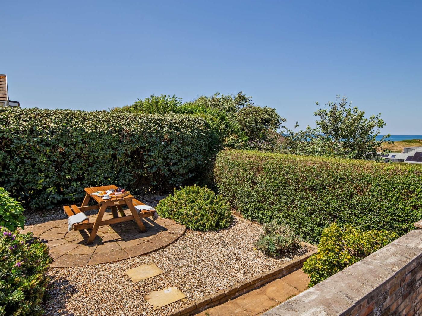 Eine Terrasse mit Holztisch und -stühlen, umgeben von Kies und niedrigen Hecken.