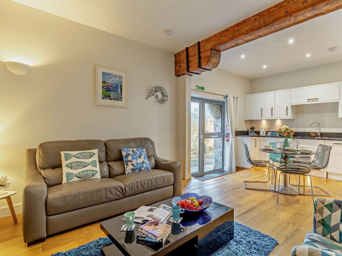 A living room with a brown sofa, a coffee table with books and a bowl of fruit, a dining area with a glass table and chairs, and a kitchen with white cabinets and a wooden beam ceiling.