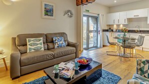 Ein Wohnzimmer mit braunem Sofa, einem Couchtisch mit Büchern und einer Obstschale, einem Essbereich mit Glastisch und Stühlen sowie einer Küche mit weißen Schränken und einer Holzbalkendecke.