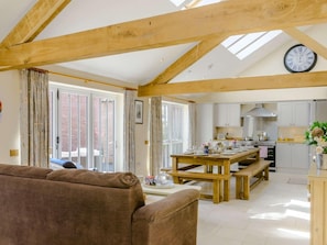 A spacious living area with a wooden ceiling, a dining table set for four, and a kitchen with white cabinets and a clock.