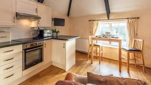 A compact kitchen with a built-in oven, a dining area with a table and chairs, and a window with curtains. - 1 Bed in Helston (oc-41377) (Cury Cross Lanes)
