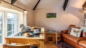 A cozy room with a wooden table set for breakfast, a leather sofa, and a painting on the wall. - 1 Bed in Helston (oc-41377) (Cury Cross Lanes)