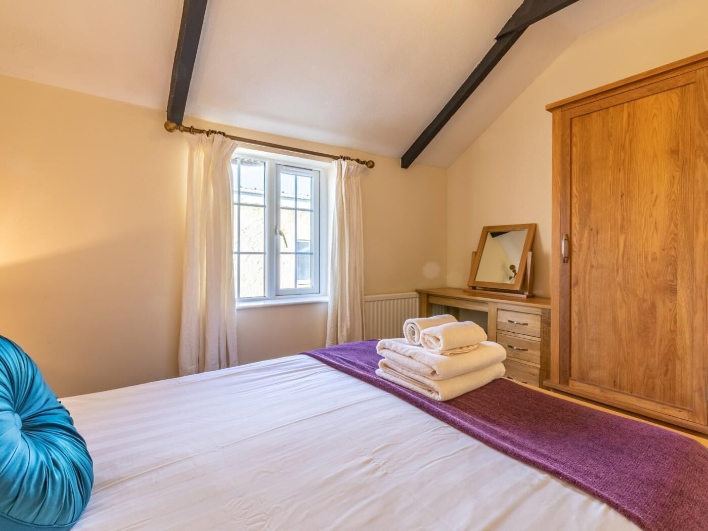 Een slaapkamer met een bed, een raam met gordijnen, een houten garderobe en een nachtkastje met een spiegel.