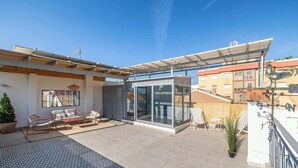 Una terraza en la azotea con cerramiento de vidrio, mobiliario exterior y una pérgola con paneles solares.
