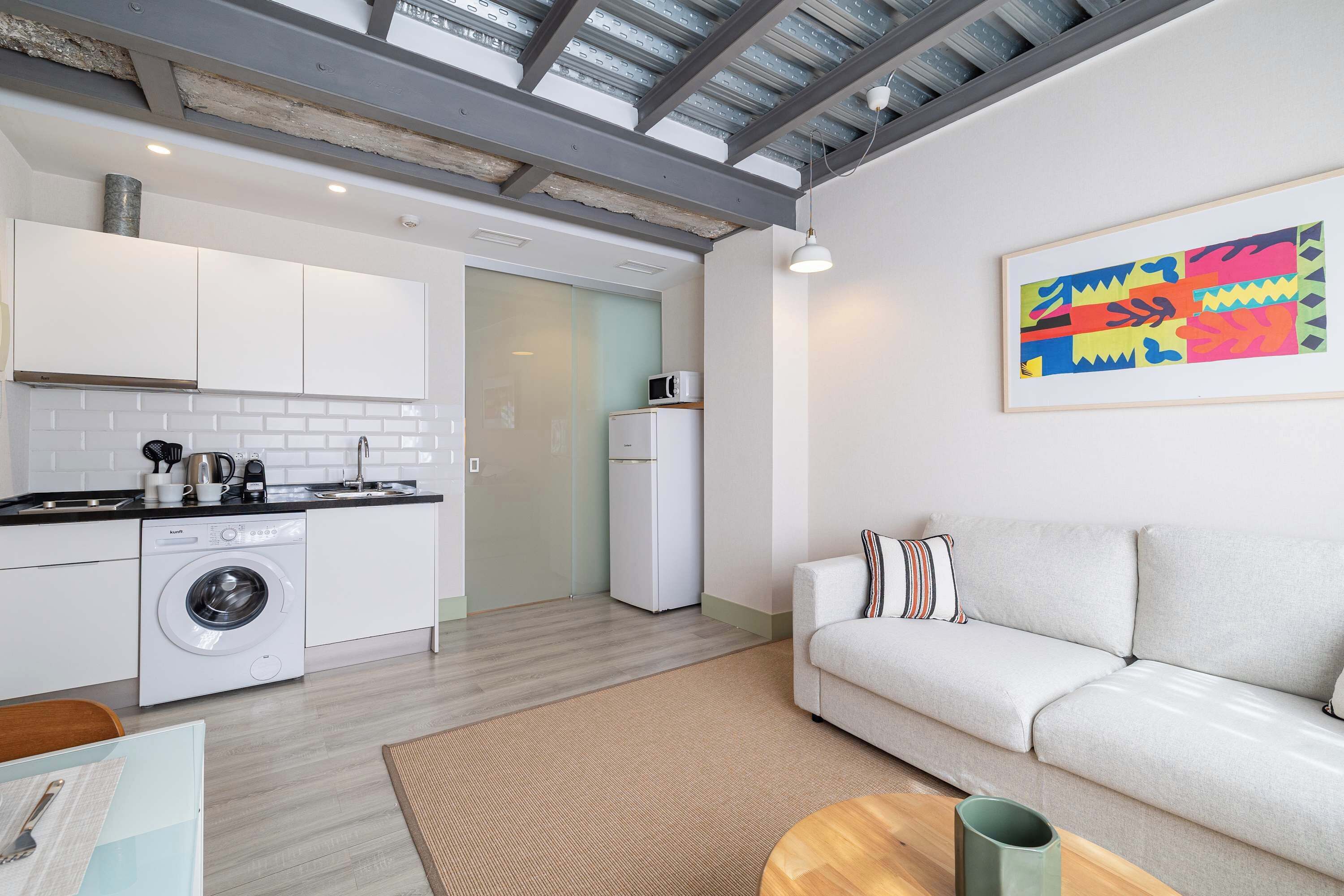 A modern apartment with a kitchen, a washing machine, a sofa, and a colorful painting.