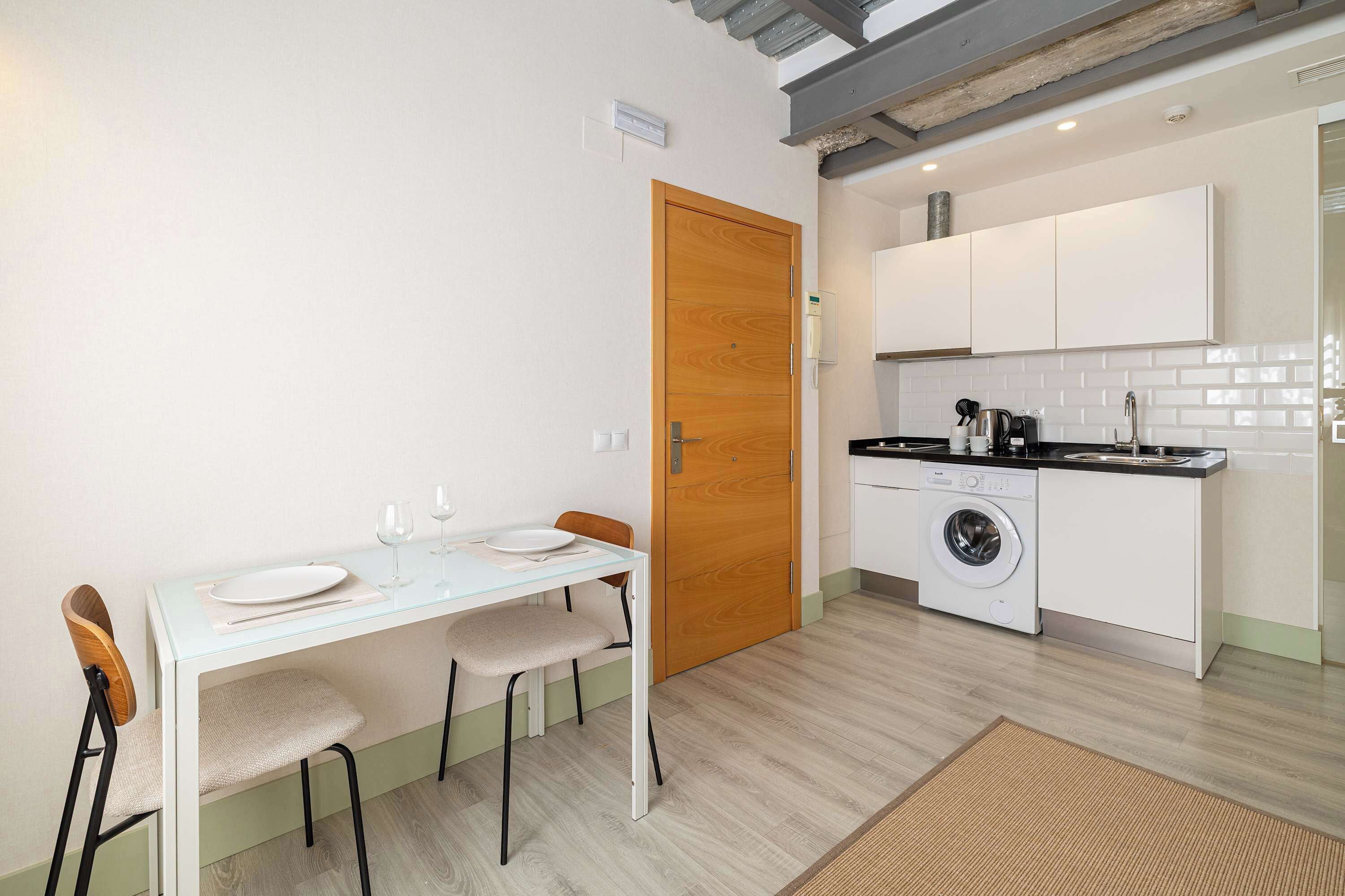Een kleine keuken met een wasmachine, een eettafel met stoelen en een houten deur.