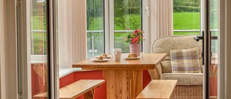 A cozy seating area with a wooden table, benches, and a wicker chair, overlooking a green landscape.