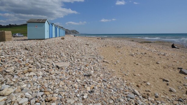 Une plage de galets bordée d’une rangée de cabanes de plage bleu clair, un ciel dégagé et une falaise lointaine.