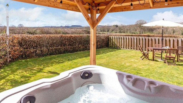 A hot tub under a wooden pergola with a view of a hedge and a table with chairs. - 2 Bed in Launceston (oc-41992) (Saint Giles on the Heath)
