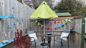 A wooden deck with a round table and two chairs, a potted plant, and a colorful triangular banner. - 1 Bed in Par- (oc-42007) (Par)