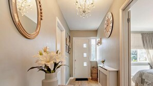 A hallway with a wooden bench, a vase of white flowers, a mirror, a clock, and a door leading to a bedroom. - 3 Bed in Ilfracombe (oc-41580) (Combe Martin)