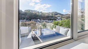 A balcony with a glass table, white cushions, and a view of a town with houses and hills. - 3 Bed in Ilfracombe (oc-41580) (Combe Martin)