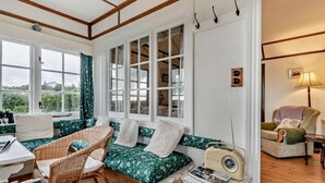 Une pièce chaleureuse avec un plancher en bois, un canapé en osier agrémenté de coussins à motifs floraux, une radio ancienne et une fenêtre avec des rideaux.