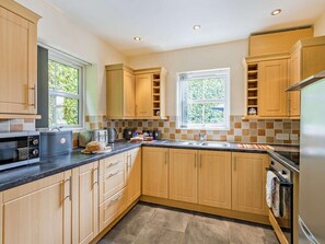 A modern kitchen with wooden cabinets, a built-in oven, a sink, and a window providing natural light. - 2 Bed in Tyn-y-gongl (oc-42369) (Marianglas)