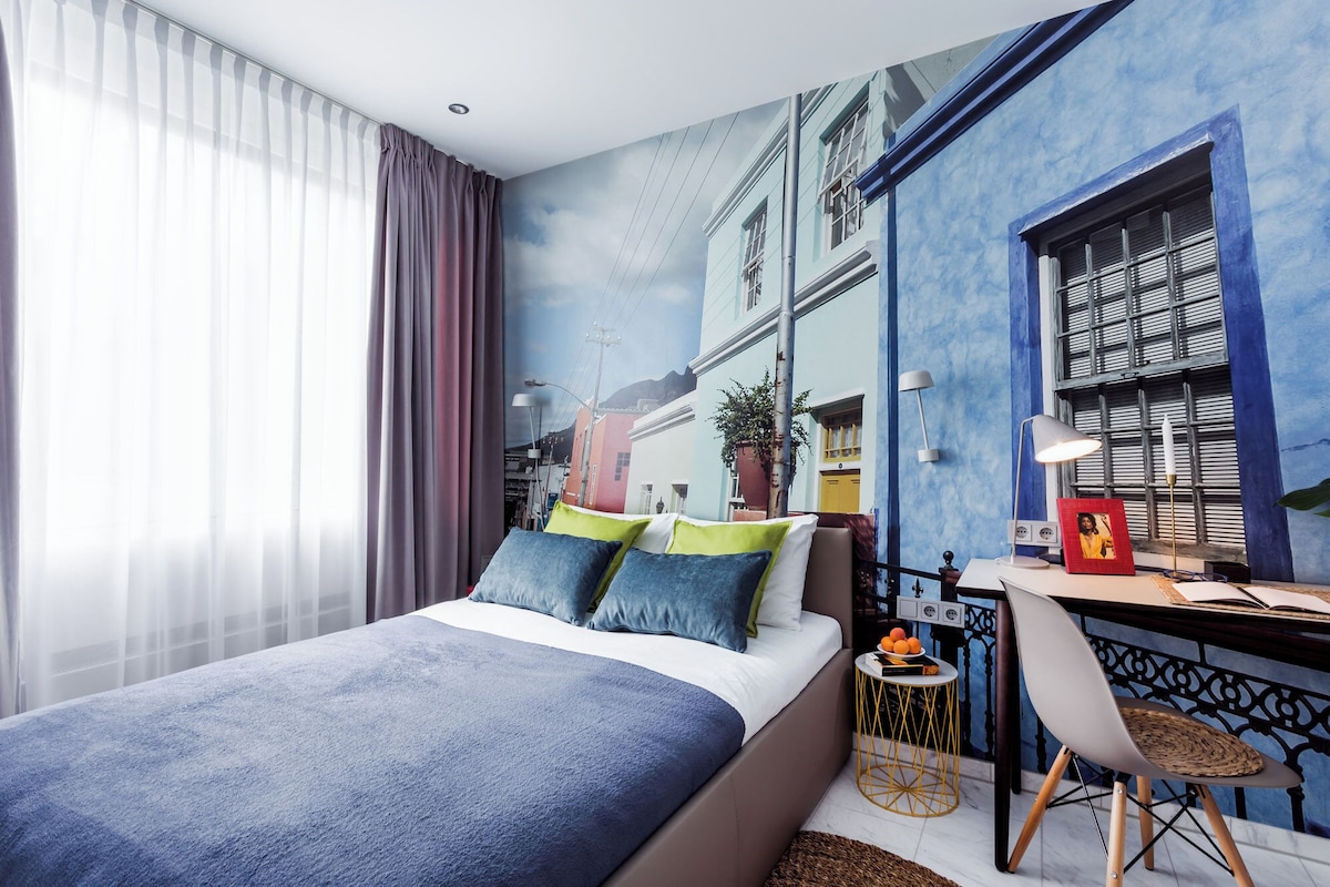 Ein Schlafzimmer mit einem Bett, einem Schreibtisch, einem Stuhl und einem Wandgemälde, das eine Straßenszene zeigt.