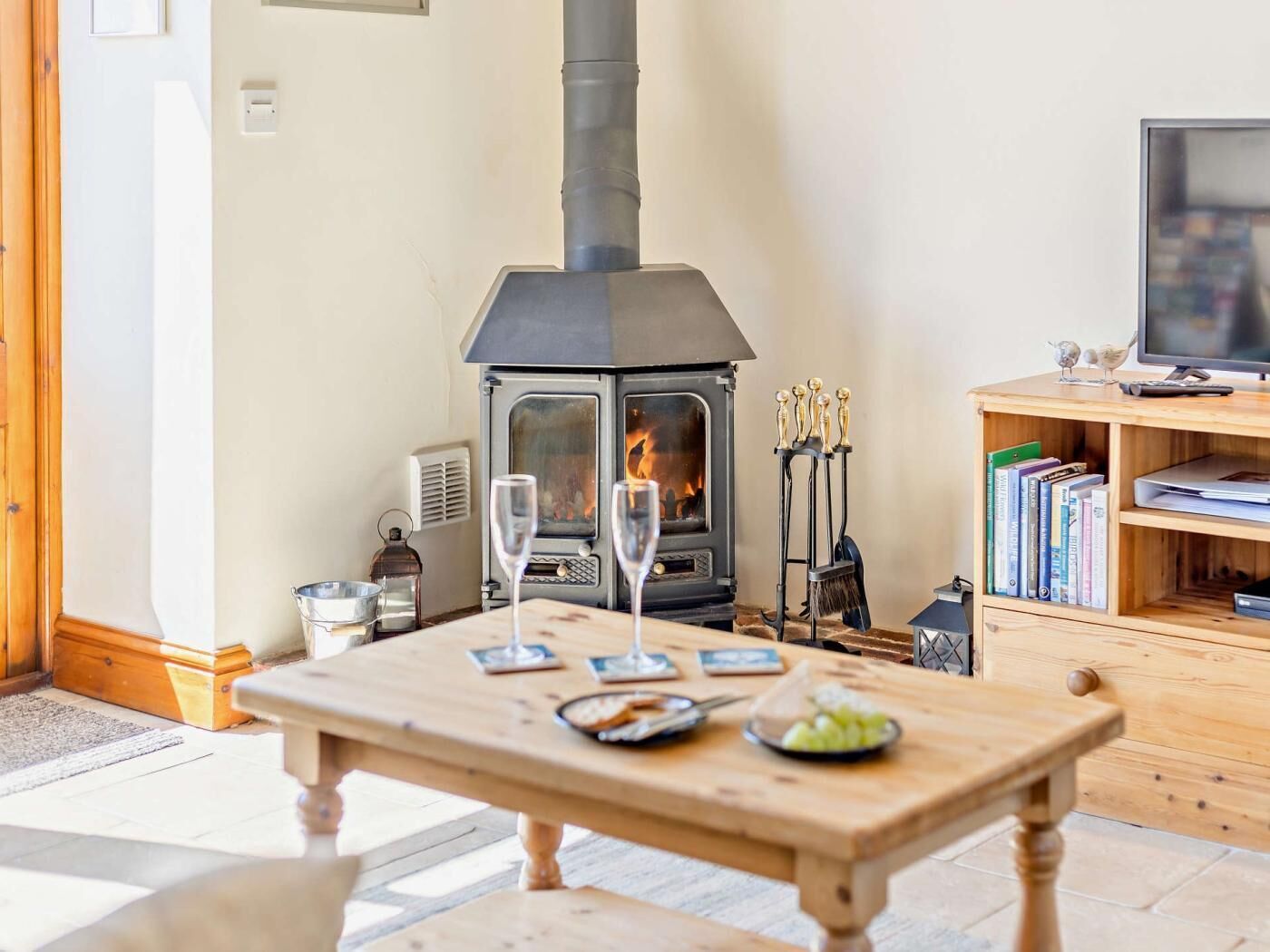 Un salón acogedor con una estufa de leña, una mesa de madera con vasos y aperitivos, una estantería y un televisor.