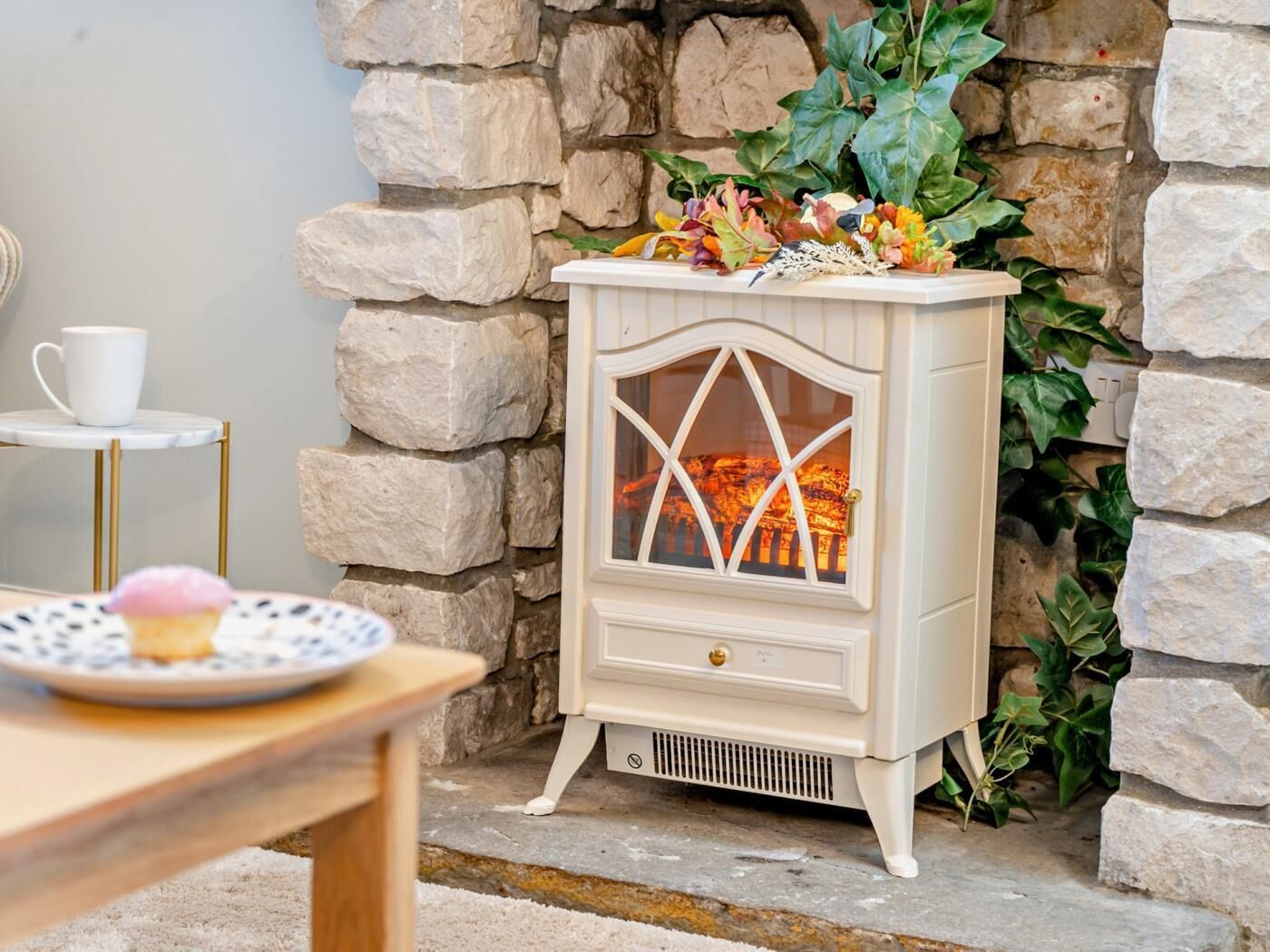 Een stenen openhaard met een decoratieve plantencompositie, een witte tafel met een bord en een kop, en een witte verwarming met een glazen deur.